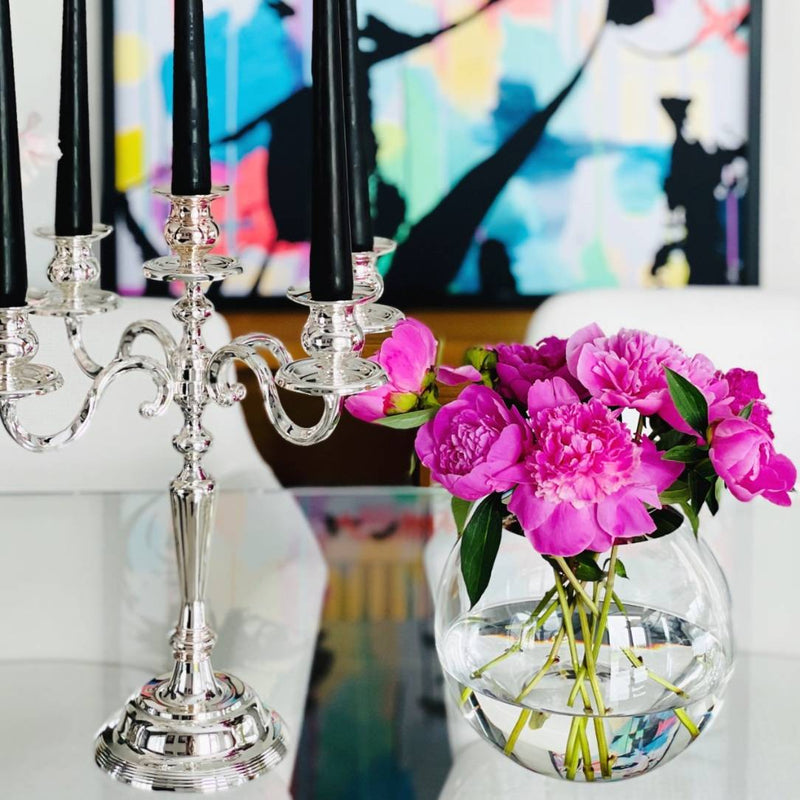 Silver candelabra with black candles and a vase of pink flowers on a table with a colorful abstract painting in the background.