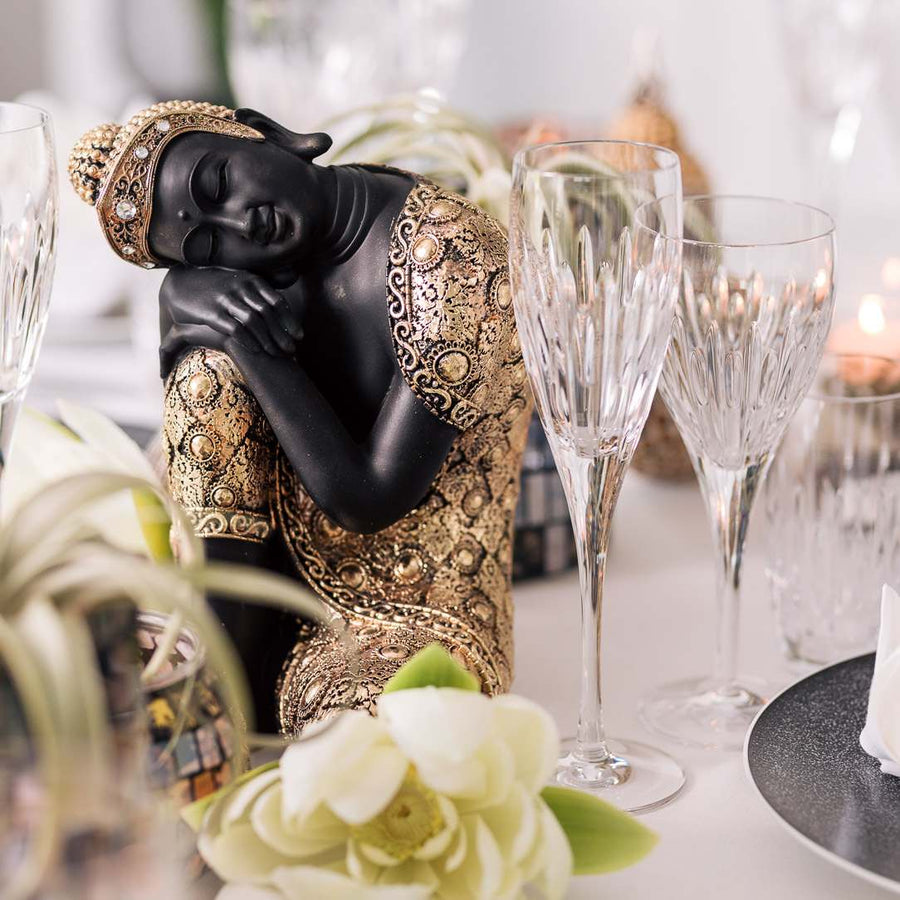 Decorative statue of a seated figure with intricate patterns on a table with glasses and flowers.