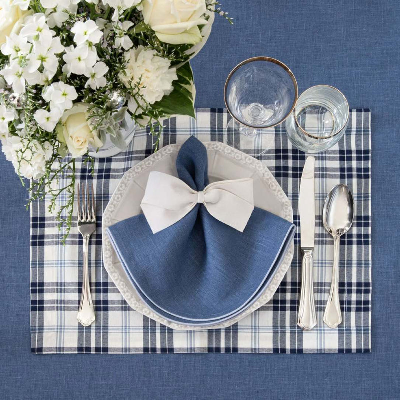 Table setting with white plates, blue napkins, silverware, and a floral arrangement on a blue plaid tablecloth.