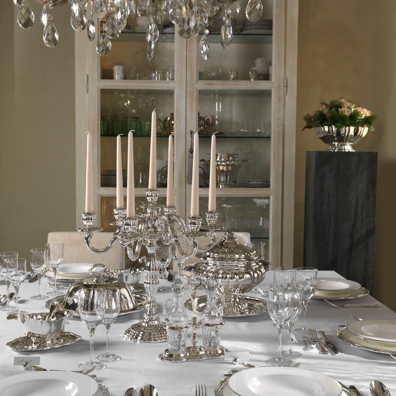 Dining table set with silverware, glasses, and a chandelier in a room with shelves.