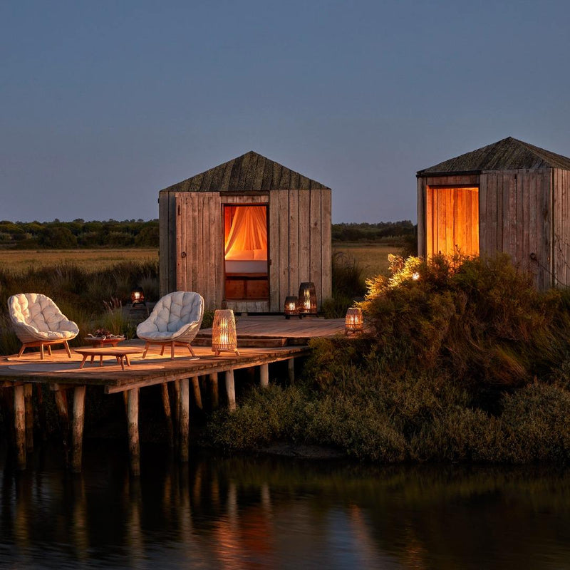 Two wooden cabins by a body of water with lit interiors and outdoor seating area.