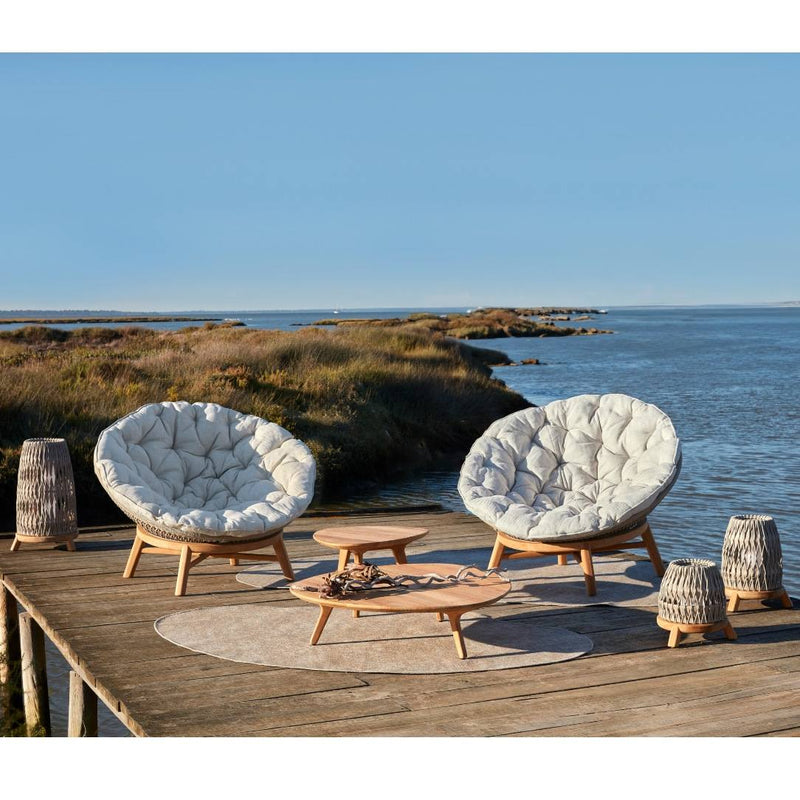 Two cushioned chairs and Sandua lanterns by Manutti on a wooden deck by the water with a clear blue sky.