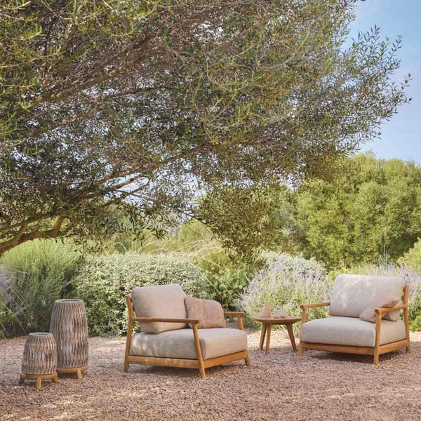 Outdoor seating area with two beige chairs, a small table and Sandua lanterns under a tree in a garden setting.