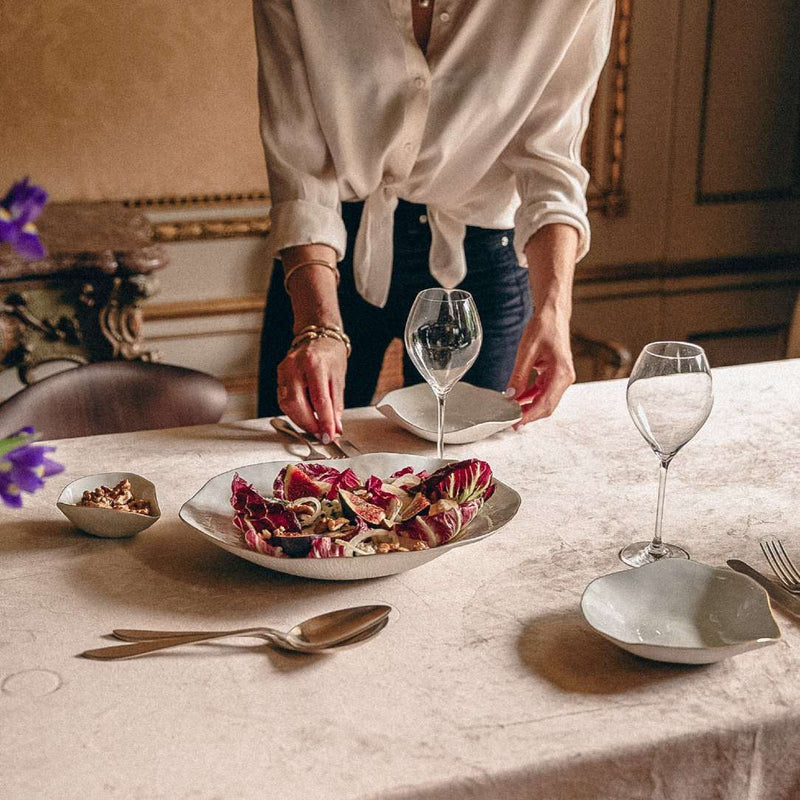 Person setting a table with elegant porcelain large dinner plate White with Golden Rim No6 Indulge Sarah-Linda  and wine glasses in a warm, indoor setting.