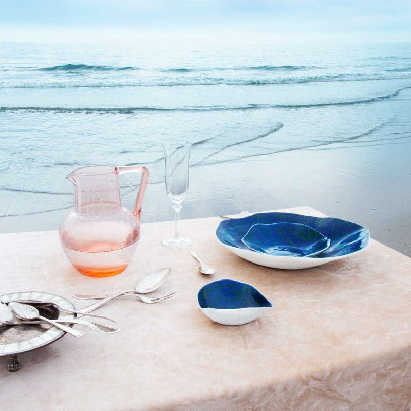 Table setting with Luxury Limoges porcelain side bowls Blue Lagoon No2 Indulge Sarah-Linda and a pink pitcher on a tablecloth against an ocean backdrop.
