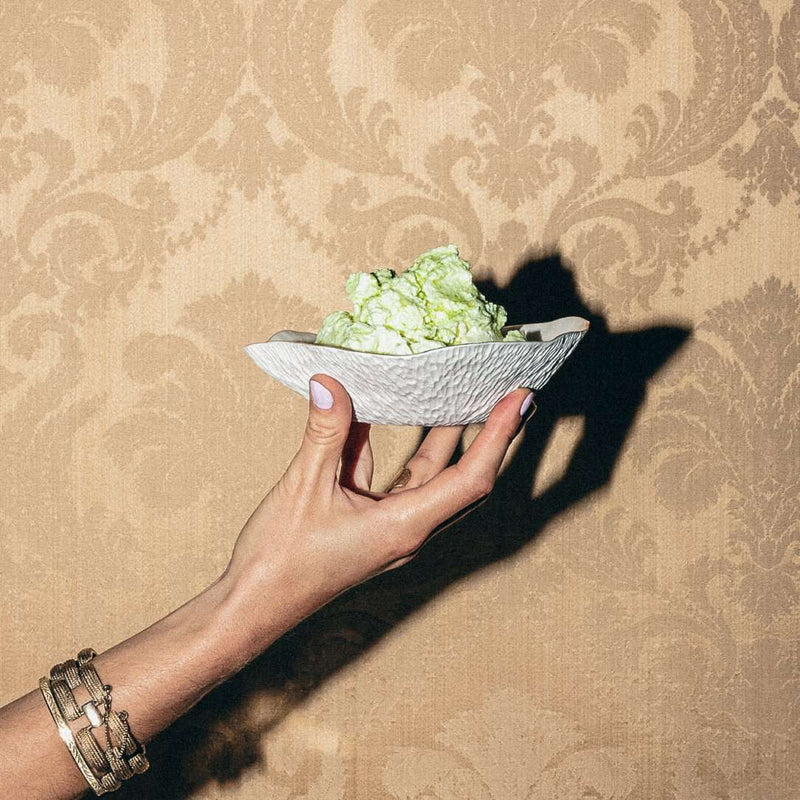 Hand holding a Shell-inspired porcelain small deep plates White with gold rim No9 Indulge with green food against a patterned beige wall.