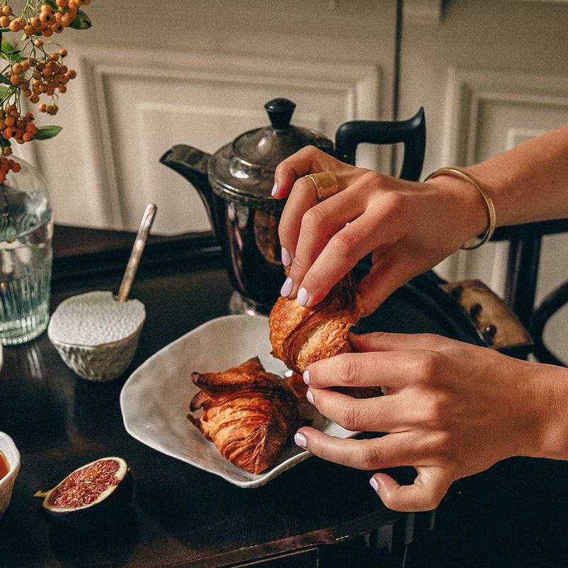 Person holding a croissant over a Shell-inspired porcelain small deep plates White finish No9 Indulge on a table with a teapot and figs.