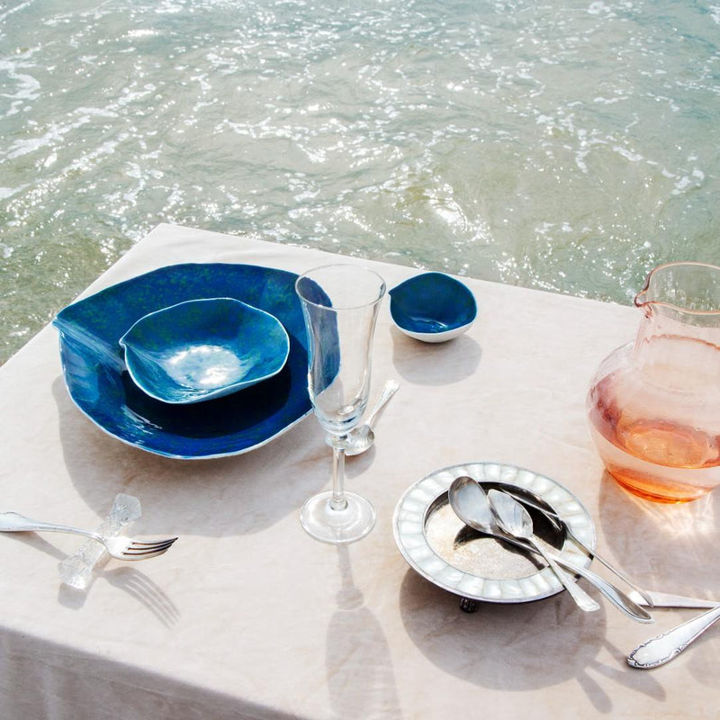 Dining table setup with Luxury Limoges porcelain small plates Blue Lagoon No5 Indulge Sarah-Linda, silverware, and a glass of water on a white tablecloth by the sea.