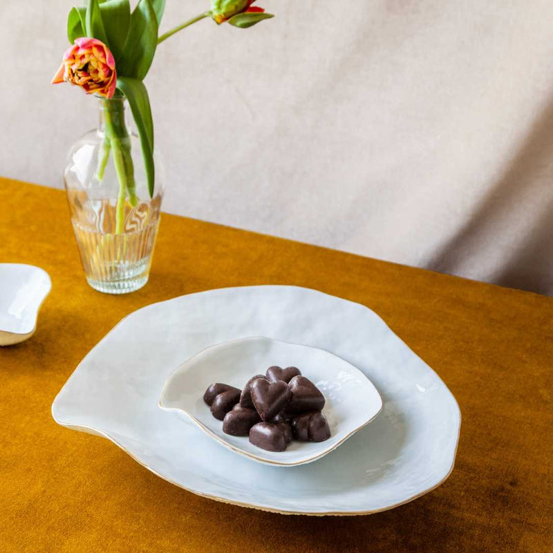 Heart-shaped chocolates on a Sculptural porcelain small plates White with Golden Rim No5 Indulge Sarah-Linda with a vase of flowers on a wooden table.