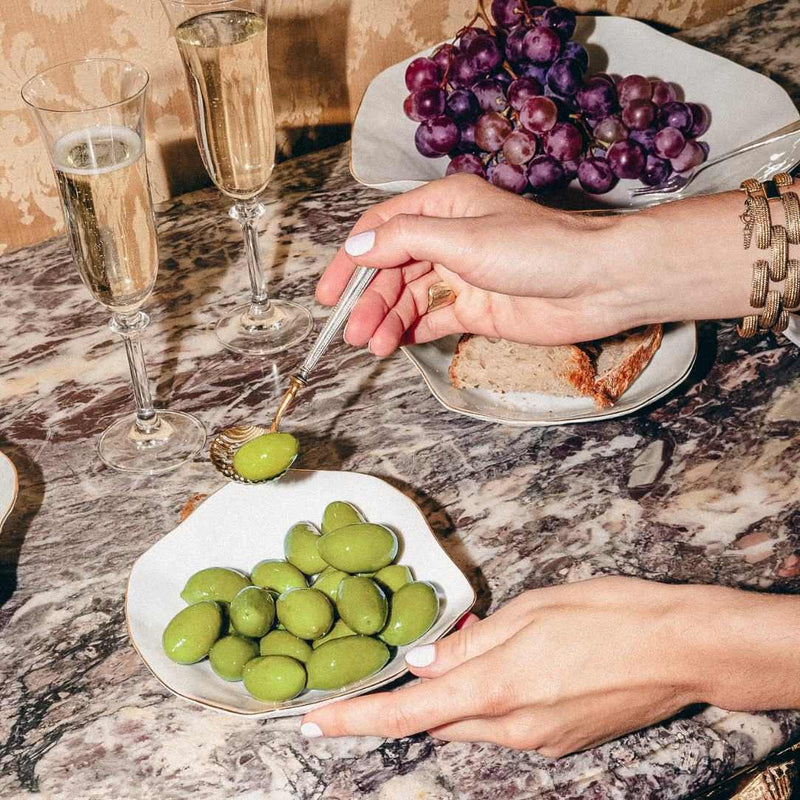 Luxury Limoges porcelain small plates White with golden rim No5 Indulge Sarah-Linda styled with olives with a spoon on a marble table with champagne and grapes.