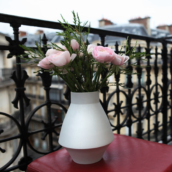 White vase with pink flowers on a red surface with a black metal railing and blurred building in the background