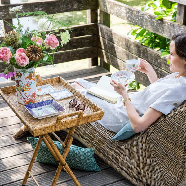Woman sitting on a wooden deck with a tray of tea and flowers, enjoying a peaceful moment.
