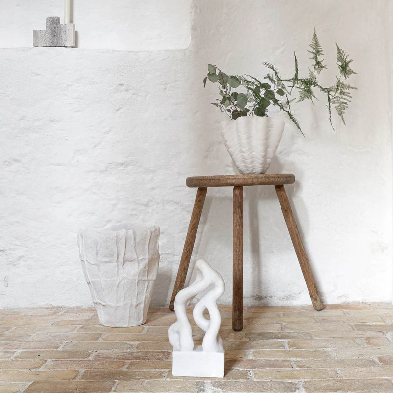 Wooden stool with a white vase and decorative plant against a white brick wall.