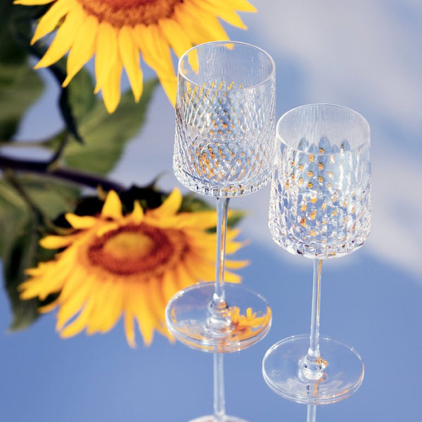 Two clear Ulysse water glass in luxury French crystal with siren-scale cuts by Cristallerie de Montbronn in front of sunflowers against a blue sky.
