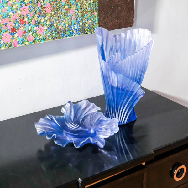 Two blue glass vases on a dark surface with a colorful floral painting in the background.