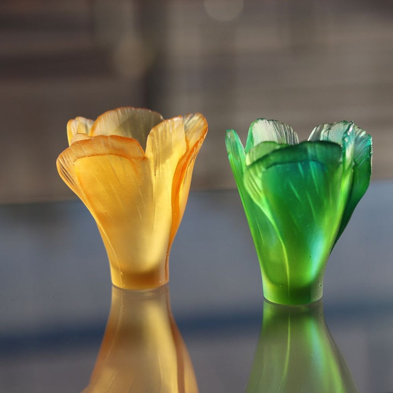 Two glass flower-shaped vases, one orange and one green, on a reflective surface with a blurred background.