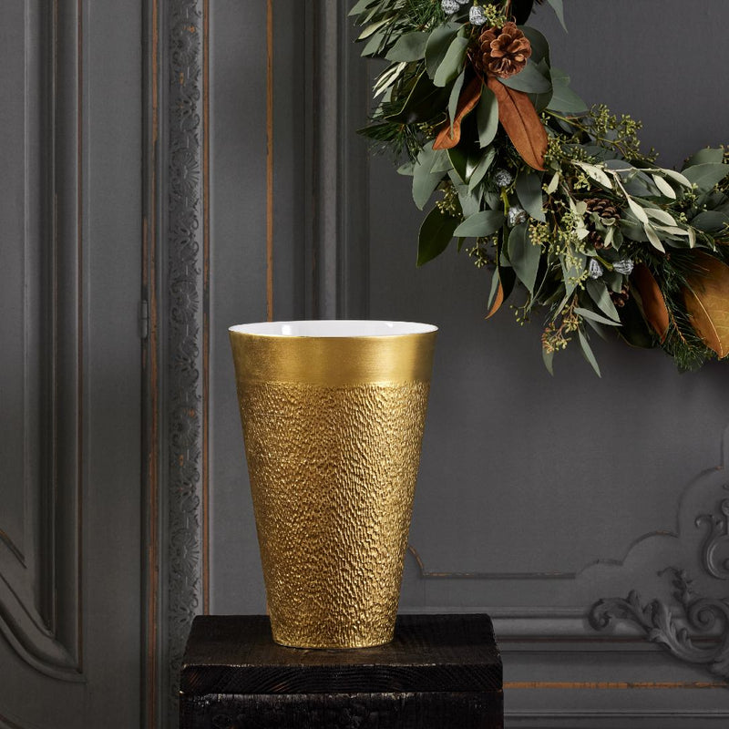 Gold textured vase on a dark surface with a decorative wreath in the background