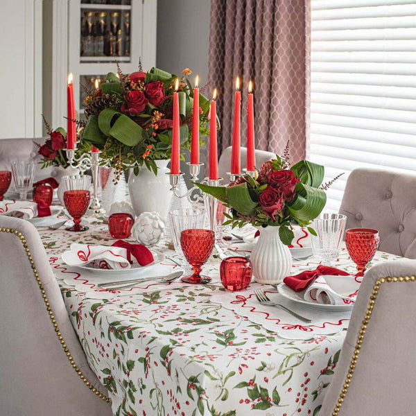 A festive Christmas-themed table setting featuring a red and green holly patterned tablecloth with red trim, accompanied by decorative red candles, white flowers, and red and clear glassware.
