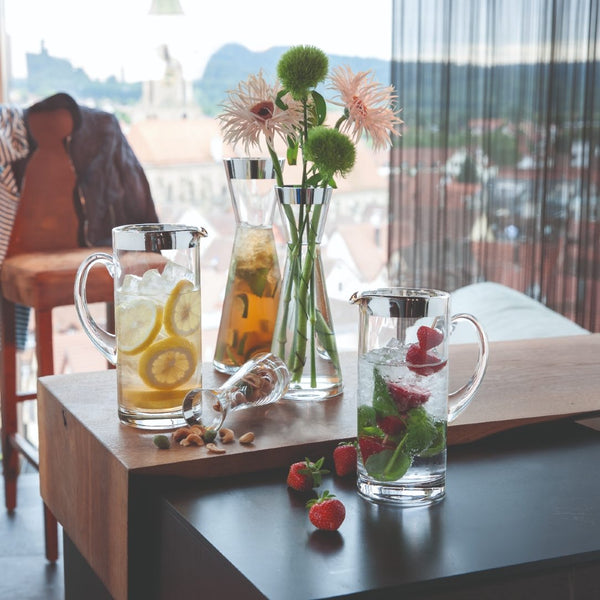 Two Elegant Handcrafted Water Jug Babsy in Crystal with Silver Rim – Sonja Quandt Glassware with iced beverages on a wooden table, surrounded by flowers and fruits.