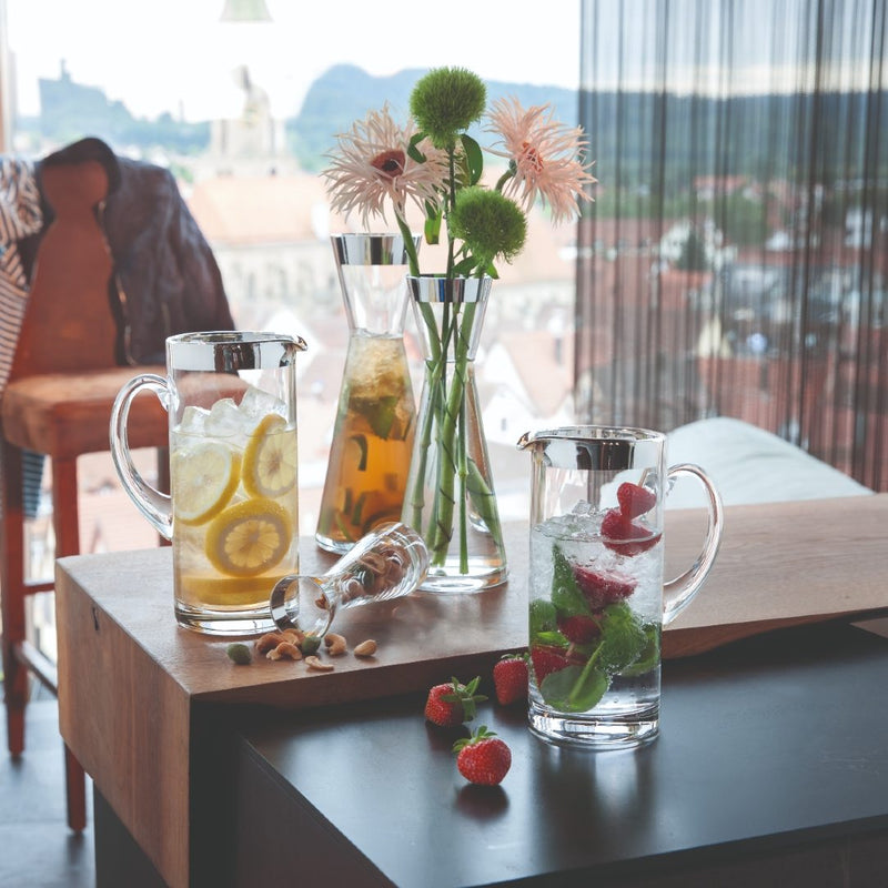 Two Elegant Handcrafted Water Jug Babsy in Crystal with Silver Rim – Sonja Quandt Glassware with iced beverages on a wooden table, surrounded by flowers and fruits.