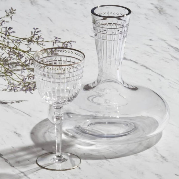 Aghata crystal wine decanter with platinum rim and geometric cut design by Cristallerie de Montbronn on a marble surface with a blurred plant in the background.