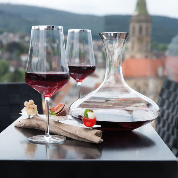 Two wine glasses with a Luxury Wine Decanter Crystal with Fine Silver Rim – Rene Collection by Sonja Quandt on a table with a scenic background.