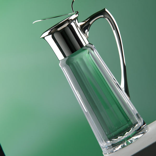 Luxury water jug Opera in sterling silver and cut crystal with hinged lid and handle by Sonja Quandt – elegant pitcher for refined dining on a green background