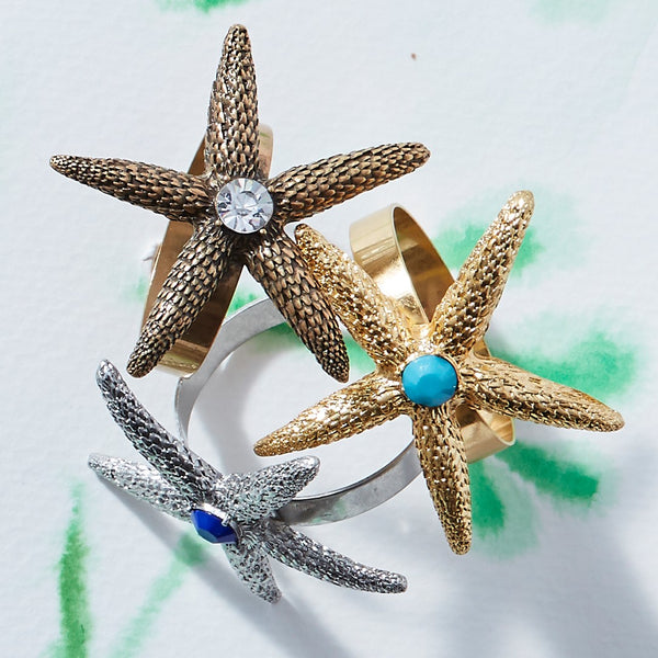 Starfish napkin rings by Joanna Buchanan in three different colors, on a white table.