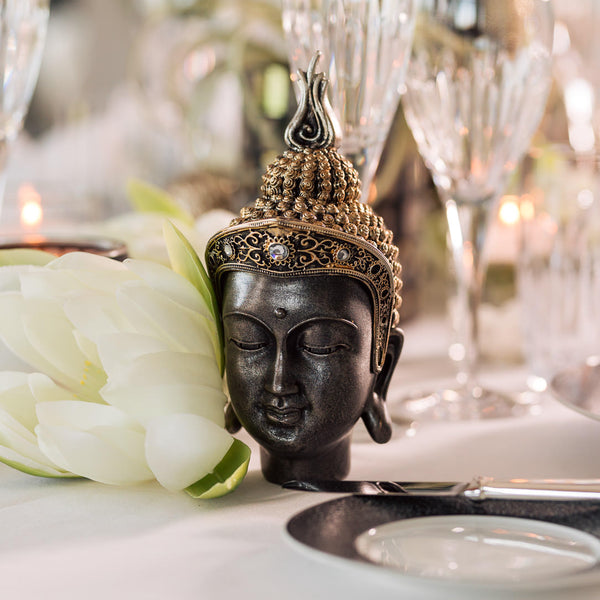 A Buddha head statue with gold and black colour, displayed on a table with lotus flower in white and green