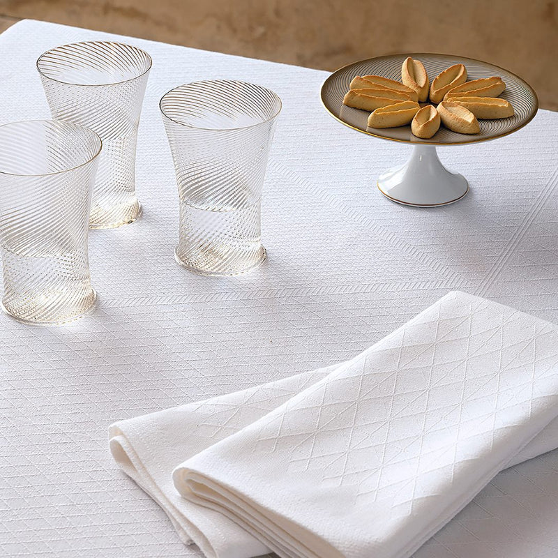 folded white napkins with geometrical pattern displayed on a white tablecloth with 3 glasses and a plate with sweets