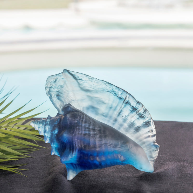 A large blue crystal glass shell sculpture designed by Daum with a sea view background.