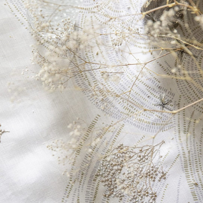Close-up of A white linen tablecloth with a large-scale embroidered design featuring coral branches, pearls, and starfish, and embroidery in beige and white gold thread.